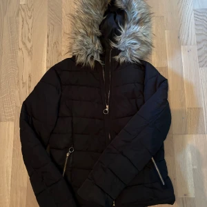 Svart pufferjacka med fuskpäls från ONLY - Svart pufferjacka från ONLY med fluffig fuskpäls runt huvan. Jackan har quiltad design, dragkedja framtill och två fickor med dragkedjor. Perfekt för kalla dagar och ger en snygg siluett. Pälsen är ljusbrun och gråtonad för extra stilpoäng.