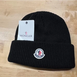 Svart ribbad mössa från Moncler - Fet svart ribbstickad mössa från Moncler med uppvikt kant och klassisk Moncler-logga framtill. Nyskick