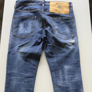 Dsquared2 blå slitna jeans med patch - Blå jeans från Dsquared2 med slitna detaljer, färgstänk och patchade hål med mönstrat tyg bakom. Klassisk femficksmodell med knappgylf och raka ben. Jeansen har en cool, edgy vibe och är tillverkade i bomull med denimkänsla. Det är riktigt feta jeans och de säljs till ett riktigt bra pris.👍storleken är 46 vilket motsvarar s/cd ungefär.