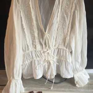 Vit blus med knut från Zara - Helt ny och använd!! Superfin vit blus från Zara i tunn bomull med v-ringning och snygga knytband framtill. Blusen har långa ärmar med volang vid ärmslut och fina spetsdetaljer längs bröstet. Råka köpa i fel storlek - men den är så så fin !! Artikelnummer på Zara 7521/242/251