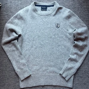 Grå stickad tröja från East West - Snygg grå stickad tröja från East West med rund halsringning och broderad patch på bröstet. Tröjan har ribbade muddar vid ärmslut och nederkant, vilket ger en clean och klassisk look. Perfekt för lager-på-lager under kyliga dagar. Skickar imon 1-2 dagar och svarar snabbt vid frågor och funderingar😄kom med prisförslag🙌