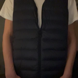 Mörkblå dunväst från Uniqlo XS - Säljer en navyfärgad, quiltad dunväst från Uniqlo i modellen Ultra Light Down. Västen är lätt, har dragkedja framtill och en stilren, enkel design utan krusiduller. Perfekt att slänga över en hoodie eller t-shirt för extra värme. Passar dig som gillar minimalistisk stil.