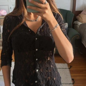 Svart spetsblus från SURE - Svart blus i spets från SURE med klassisk krage och vita knappar framtill. Genomskinligt spetsmaterial med blommigt mönster och lång ärm. Snygg, feminin passform som sitter normal. Perfekt för dig som vill ha en cool och elegant look.