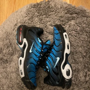 Nike Air Max Plus blå/svarta sneakers - Nike Air Max Plus sneakers i blått och svart med vita detaljer. Skorna har mesh-överdel, svarta vågformade linjer och tydlig Air Max-sula. Swoosh-logga på sidan och Tn-logo bak. Skorna har snörning och en sportig vibe, perfekta för dig som gillar streetwear.