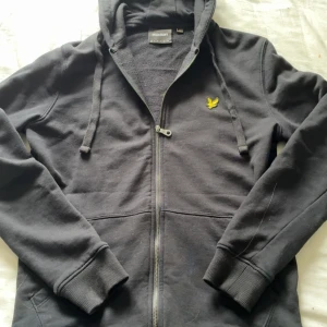 Svart hoodie från Lyle & Scott - Svart hoodie från Lyle & Scott med dragkedja och huva. Tröjan har snörning i huvan, ribbade muddar och den klassiska gula loggan broderad på bröstet. Tillverkad i mjuk bomull, perfekt för chill dagar.
