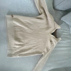 Beige stickad tröja Polo Ralph Lauren - Snygg beige stickad tröja från Polo Ralph Lauren med diskret broderad logga på bröstet. Tröjan har långärmad design, ribbade muddar och rund hals med liten v-ringad detalj. Perfekt för dig som gillar klassisk och clean stil.
