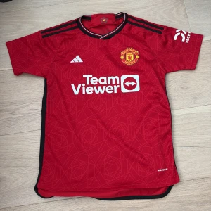 Manchester United Adidas matchtröja S - Röd Manchester United matchtröja från Adidas med geometriskt mönster, klubbmärke på bröstet och TeamViewer-tryck. Svarta och vita detaljer på krage och axlar. Tillverkad i lätt och ventilerande polyester, perfekt för fotboll eller supporterliv.