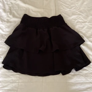 Svart volangkjol från ONLY, storlek 38 - Supersöt svart kortkjol från ONLY med bred smockad midja och dubbla volanger. Kjolen har en loose passform och är perfekt för dig som gillar en enkel men trendig look. Materialet känns mjukt och lätt, vilket gör den bekväm att bära hela dagen.