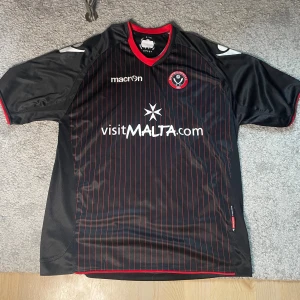 Sheffield United fotbollströja 2010/11 - Sheffield United FC fotbollströja från Macron i svart med tunna röda ränder. Kortärmad modell med röd krage och klubbmärke på bröstet. Tillverkad i lätt och ventilerande polyester, perfekt för match eller träning. Snygg sponsorlogga och detaljer på ärmarna.