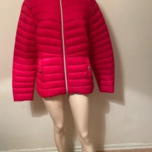 Rosa pufferjacka med dragkedja - Snygg pufferjacka i stark röd färg med horisontella sömmar och två stora fickor framtill. Jackan har en silvrig dragkedja och tryckknappar på fickorna. Perfekt för kyliga dagar och ger en cool vibe till din outfit.