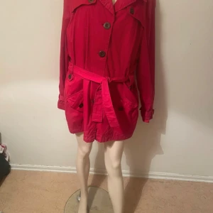 Röd trenchcoat med bälte och knappar - Snygg röd trenchcoat med klassisk krage, stora knappar och bälte i midjan. Jackan har två stora fickor framtill och långa ärmar med knäppning vid ärmslut. Perfekt för dig som vill sticka ut med färg i vår eller höst storlek 44 men passar 46 också 