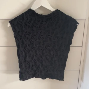 Svart ärmlös topp med strukturmönster - Säljer en svart ärmlös topp med coolt strukturmönster som ger en unik look. Toppen har rund hals och är tillverkad i ett mjukt material som känns skönt mot huden. Perfekt att styla med jeans eller kjol för en trendig outfit.