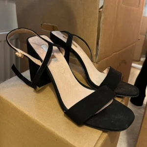 Svarta sandaler med blockklack - Stilrena svarta sandaler med bred blockklack och öppen tå. Skorna har en enkel rem över tårna och en smal rem runt hälen med guldfärgat spänne. Perfekta för att lyfta din outfit med en modern touch.