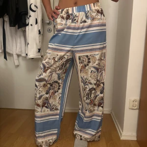 Blommiga vida byxor med blå ränder - Säljer ett par superbekväma vida byxor med blommigt mönster och breda blå och vita ränder. Byxorna har resår i midjan och är gjorda i ett lätt material som känns luftigt. Perfekta för dig som gillar en chill och snygg stil.