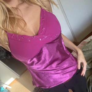 Rosa satinlinne från Secret Possessions - Säljer ett glansigt rosa linne från Secret Possessions med tunna axelband och broderade blommor längs v-ringningen. Toppen har en snygg satinfinish och är perfekt för dig som gillar färg och detaljer. Passar dig som vill sticka ut lite extra.