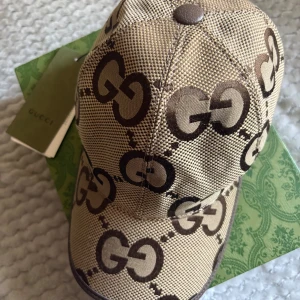 Gucci Keps - Snygg beige keps från Gucci med stora bruna GG-loggor över hela kepsen. Tillverkad i 100% bomull och har justerbar rem baktill i brunt skinn med präglad Gucci-text. Perfekt accessoar för att lyfta din outfit.