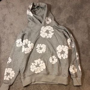 Grå hoodie från Denim Tears med blommor - Grå hoodie från Denim Tears med stora vita blommönster över hela tröjan. Tröjan har huva och ribbade muddar vid ärmslut och nederkant. Perfekt för dig som vill sticka ut med en unik streetwear-look.