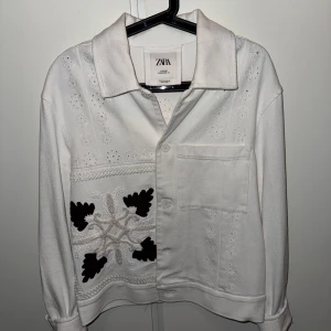 Vit broderad overshirt från Zara - Säljer en vit overshirt från Zara med broderade detaljer och svart brodyr framtill. Jackan har en bröstficka, knäppning framtill och krage. Materialet är bomull och plagget har lång ärm. Perfekt för dig som gillar unika detaljer och stilren look.
