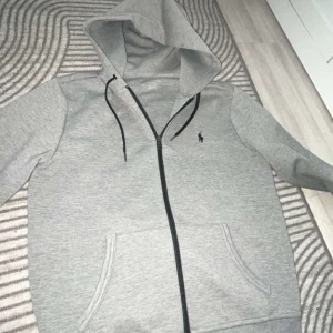 Grå hoodie från Ralph Lauren - Grå hoodie från Ralph Lauren med dragkedja och svart logga på bröstet. Tröjan har huva med svarta snören och två fickor framtill. Perfekt för en chill och avslappnad stil.