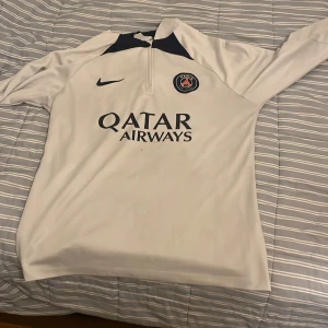 PSG långärmad träningströja Nike M - Säljer en långärmad Paris Saint-Germain träningströja från Nike i Dri-FIT-material. Tröjan är vit med marinblå detaljer, halv dragkedja och PSG-logga samt Qatar Airways-tryck på bröstet. Perfekt för fotbollsträning eller supporterstil.