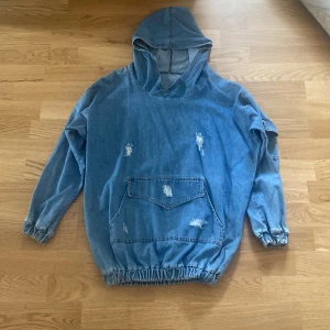 Oversized jeanshoodie med huva - Säljer en oversized hoodie i ljusblå denim med huva och stora fickor framtill. Tröjan har slitna detaljer, resår i midjan och vid ärmsluten samt lång ärm med extra ficka på ena ärmen. Perfekt för en avslappnad streetstyle-look.