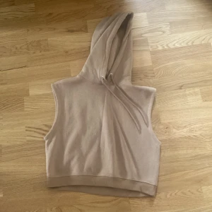 Beige croppad hoodie utan ärmar - Säljer en beige croppad hoodie utan ärmar med huva och snörning. Perfekt för lager-på-lager och chill dagar. Mjuk och skön i bomullsmix, enkel att matcha med jeans eller tights.