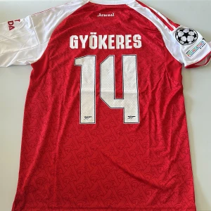 Arsenal Gyökeres #14 matchtröja Adidas - Röd och vit Arsenal fotbollströja från Adidas med Gyökeres och nummer 14 på ryggen. Tröjan har korta ärmar, Champions League-märke på ärmen och diskret mönster i tyget. Broderat klubbmärke och sponsorlogga framtill. Tillverkad i lätt funktionsmaterial. Replika, endast handtvätt gäller!
