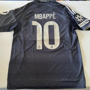 Real Madrid Mbappé #10 bortatröja Adidas - Snygg Real Madrid bortatröja med Mbappé och nummer 10 på ryggen. Tröjan är mörkblå med tunna ränder, vita detaljer och gula inslag. Broderade klubbmärken, Champions League-märke och 'Emirates Fly Better' på bröstet. Tillverkad i lätt och ventilerande polyester. Replika, endast handtvätt gäller!