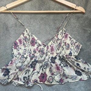 Blommig linnetopp från Zara i S - Supersöt linnetopp från Zara med blommigt mönster i lila, svart och beige. Toppen har smala axelband, v-ringning och volangig nederdel. Materialet är lätt och luftigt, perfekt för varma dagar. Knytning i ryggen för extra detalj. Säljs i storlek S. Den är väldigt efterfrågad där av priset osäker om ja vill sälja bara om ja får ett bra pris 
