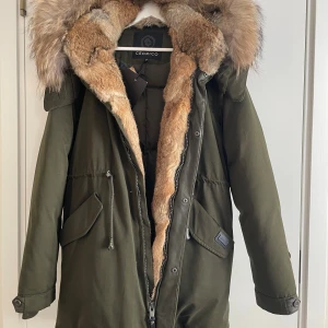Olivgrön parkas från Cedrico med päls - Snygg olivgrön parkas från Cedrico med stor fluffig äkta päls runt huvan och insidan. Jackan har dragkedja, knappar, snörning i midjan och två stora fickor framtill. Perfekt för kalla dagar med sin vadderade insida och exklusiva känsla. Jackan har aldrig använts då prislappen och alla etiketter sitter kvar. 