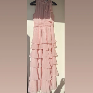 Ljusrosa volangklänning från Neo Noir - Supersöt långklänning från Neo Noir – ljusrosa Jag råkade beställa fel storlek, den är tyvärr för stor för mig. Klänningen är perfekt för bal, bröllop eller andra sommarfester! 	•	Ärmlös med volanger i flera lager 	•	Fin knytning vid axeln 	•	Tillverkad i lätt och luftigt material för en romantisk känsla 	•	Färg: ljusrosa  En vacker och elegant klänning som passar alla sommarens festligheter!🌸🎀