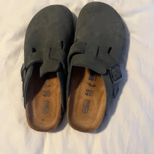 Birkenstock Boston mörkgrå mocka - Säljer ett par Birkenstock Boston i mörkgrå mocka med justerbart spänne i metall. Skorna har en klassisk rund tå och platt sula med mönstrad undersida. Perfekta för dig som gillar bekväma och stilrena sandaler med en snygg och enkel design. Nypris 1800 dessa är knappt använda. Använda 3 gånger i skolan men tyvärr för små för mig och rensar garderoben så priset är inte hugget i sten så kom gärna med prisförslag.