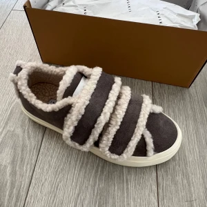 Sneakers från Inuikii - Unika sneakers från Inuikii i grå mocka med fluffiga detaljer i beige fårull. Skorna har tre breda kardborreband och en vit platt sula. Insidan är fodrad med mjuk fårull för extra komfort och värme. Perfekta för dig som vill ha något både snyggt och mysigt.