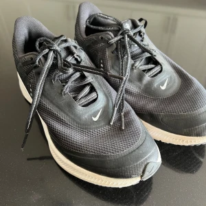Svarta Nike sneakers strl 38 - Säljer ett par svarta Nike sneakers i storlek 38 med vit sula och klassisk snörning. Ovandelen är i mesh och syntetmaterial för bra andningsförmåga. Snygg, sportig look med diskreta vita detaljer och Nike-logga på sidan och tungan.