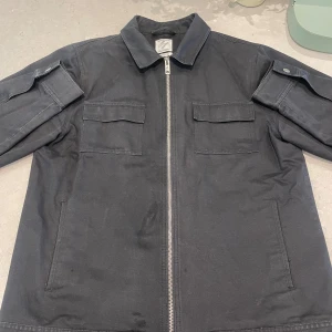 Svart overshirt med dragkedja - Stilren svart overshirt från ClubsAndSpades med raka linjer, två bröstfickor med lock, sidofickor och silverfärgad dragkedja. Jackan har klassisk krage och ärmar med knappdetalj. Perfekt för lager-på-lager och en clean look.