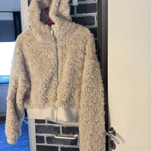 Beige teddyjacka med huva och dragkedja - Mysig beige teddyjacka med fluffig yta, huva och hel dragkedja framtill. Jackan har ribbade muddar och midja för en skön passform. Perfekt för kyliga dagar när du vill ha en chill och trendig look. Aldrig använd, prislappen på. 