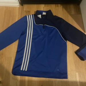 Blå Adidas half zip träningströja - Säljer en blå och marinblå långärmad träningströja från Adidas med klassiska vita ränder på framsidan. Tröjan har en hög krage med half zip ,perfekt för sport eller fotbollsträning.