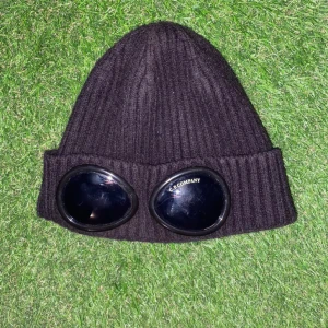 Cp Company mössa  - Svart ribbstickad mössa från C.P. Company med ikoniska svarta goggles framtill på uppviket. Mössan har en klassisk form och är tillverkad i mjukt ullmaterial. Perfekt för dig som vill sticka ut med streetwear-vibe och snygg detalj.
