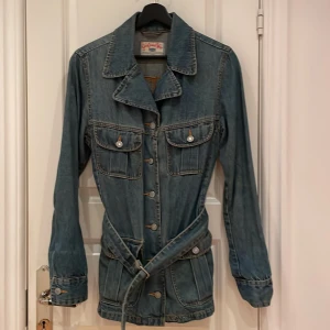 Jeansjacka med bälte från Rocky - Snygg blå jeansjacka i sliten look från Rocky med markerad krage, fyra fickor framtill och bälte i midjan. Jackan har knappar hela vägen och långa ärmar. Perfekt för dig som gillar en klassisk denimlook med lite extra detaljer.