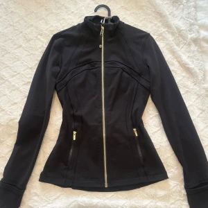 Svart träningsjacka från Lululemon - Snygg svart träningsjacka från Lululemon med figurnära passform, hel dragkedja i guld och två fickor med dragkedja. 