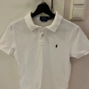 Vit pikétröja Polo Ralph Lauren - Vit pikétröja från Polo Ralph Lauren i slim fit-modell. Klassisk krage, korta ärmar och knäppning framtill. Ikonisk broderad logga på bröstet. Tillverkad i bomull för en skön och luftig känsla. Storleken är 14-16 år, samma som XS/S