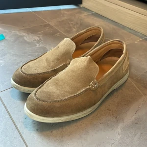 Beige mockaloafers med vit sula - Snygga loafers i beige mocka med vit sula och diskreta sömmar. Skorna har en klassisk, rund tå och är perfekta för en avslappnad men stilren look. Passar till både jeans och chinos för en chill vibe.