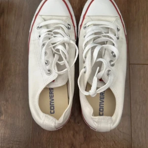 Vita Converse sneakers strl 38 - Klassiska vita Converse sneakers med lågt skaft, röda detaljer och vita snören. Skorna har rund tå, platt sula och är tillverkade i textil. Perfekta för en avslappnad och cool stil. Unisexmodell i storlek 38.