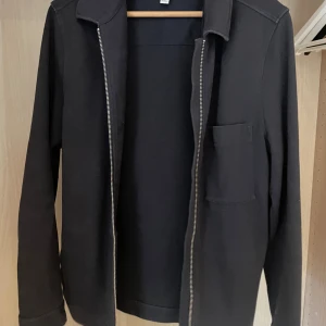 Svart overshirt med dragkedja - Stilren svart overshirt med dragkedja framtill och en bröstficka. Jackan har klassisk krage och raka ärmar, perfekt för lager-på-lager. Enkel och clean design som funkar till det mesta.