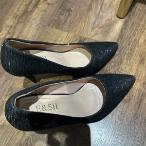 Svarta glittriga pumps med spetsig tå - Snygga svarta pumps från E&SH med glittrig yta och spetsig tå. Skorna har hög klack och en elegant form som ger en stilren look. Perfekta för dig som vill sticka ut med lite extra glitter. Storlek 37.