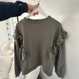 Olivgrön sweatshirt med volang från Gina Tricot - Olivgrön sweatshirt från Gina Tricot med långa ärmar och snygga volangdetaljer längs axlar och ärmar. Tröjan har rund halsringning och är gjord i mjuk bomull, perfekt för en chill och trendig look.