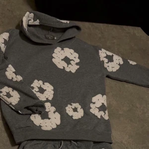 Denim Tears Set - Grå hoodie med stora vita blommönster över hela tröjan. Tröjan har huva och långa ärmar, perfekt för en chill och avslappnad stil. Mjuk och skön, passar till både jeans och mjukisbyxor. DENIM TEARS.