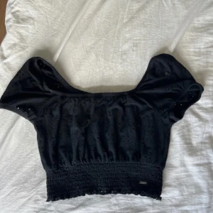 Svart croppad topp från Hollister - Supersöt svart croppad topp från Hollister med puffiga korta ärmar och smockad midja. Toppen har broderade hål och elastisk halsringning som kan bäras offshoulder. Perfekt för en trendig och chill look.