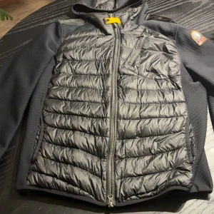 Svart hybridjacka från Parajumpers - Snygg svart hybridjacka från Parajumpers med quiltad framsida och rygg i glansigt material, samt ärmar och sidor i stretchig mesh. Jackan har huva, dragkedja framtill, bröstficka och logga på ärmen. Perfekt för kyliga dagar. Ops den har en defekt på baksidan men den syns knappt har fixat den 