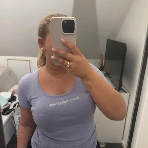 Ljuslila t-shirt från Emporio Armani - Snygg ljuslila t-shirt från Emporio Armani med rund halsringning och korta ärmar. T-shirten har ett glittrigt Emporio Armani-tryck framtill och är gjord i mjuk bomull som sitter skönt mot kroppen. Perfekt för dig som gillar stilrena och trendiga plagg. Toppen är i mycket fint skick, några stenar har gått av men inget som syns eller märks av.